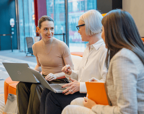 Three women sitting on a couch, each using a laptop, engaged in conversation and collaboration.